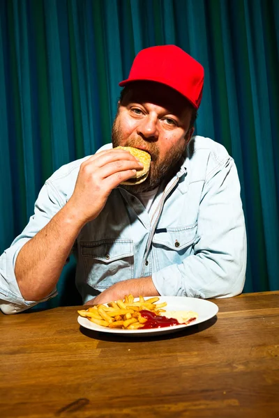 吃快餐餐大胡子的男人。享受法式炸薯条和汉堡。卡车司机与红章. — 图库照片