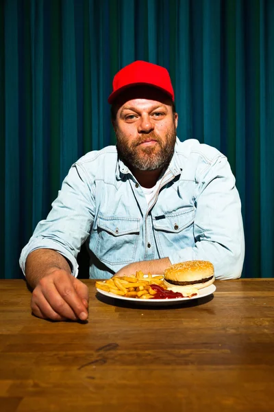 Man met baard eten van fast-food maaltijd. genieten van Franse frietjes en een hamburger. Trucker met rode dop. — Stockfoto