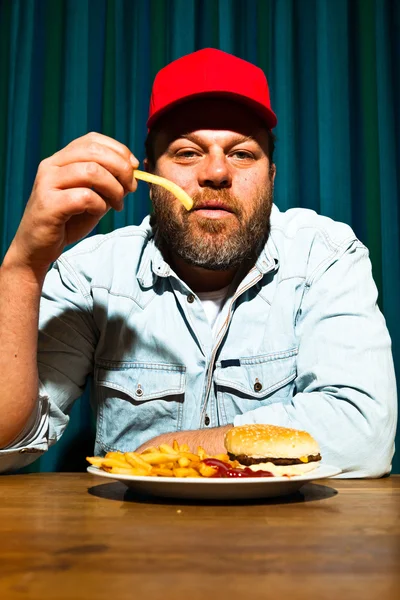 Un uomo con la barba che mangia fast food. Mi godo patatine fritte e un hamburger. Camionista con cappuccio rosso . — Foto Stock