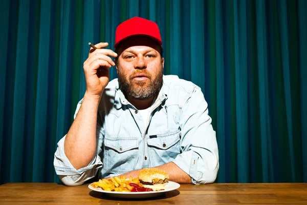 Mannen med skägget äter snabbmat måltid. njuter av pommes frites och en hamburgare. röka en cigarr. Trucker med röd mössa. — Stockfoto