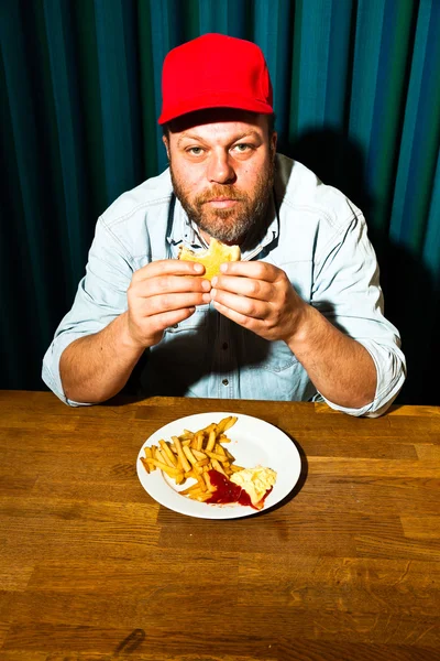 Άνθρωπος με γένια τρώνε γεύμα fast food. απολαμβάνοντας τις πατάτες τηγανιτές και ένα χάμπουργκερ. φορτηγατζή με το κόκκινο σκουφί. — Φωτογραφία Αρχείου