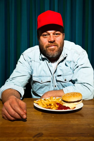 Férfi szakállal eszik gyorséttermi étkezés. élvezi a sült krumpli és a hamburger. traktoros piros sapkát. — Stock Fotó