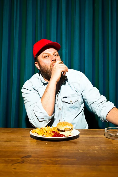 Homem com barba a comer comida rápida. A desfrutar de batatas fritas e de um hambúrguer. Fumar um charuto. Camionista com tampa vermelha . — Fotografia de Stock