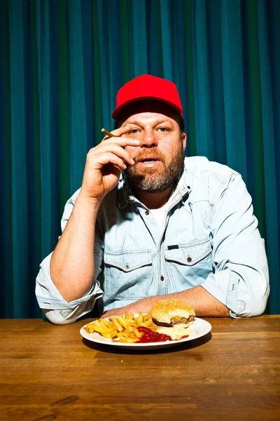 Hombre con barba comiendo comida rápida. Disfrutando de papas fritas y una hamburguesa. Fumar un cigarro. Camionero con gorra roja . — Foto de Stock
