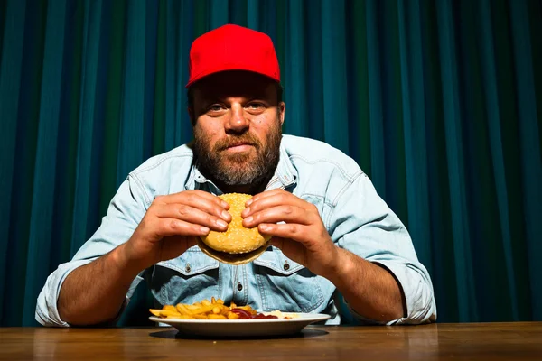 Férfi szakállal eszik gyorséttermi étkezés. élvezi a sült krumpli és a hamburger. traktoros piros sapkát. — Stock Fotó