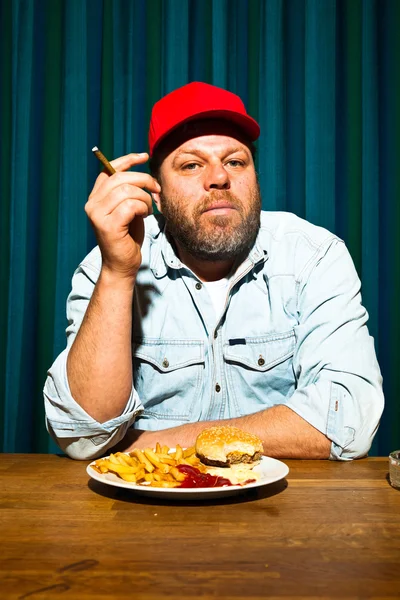 Un uomo con la barba che mangia fast food. Mi godo patatine fritte e un hamburger. Fumo un sigaro. Camionista con cappuccio rosso . — Foto Stock