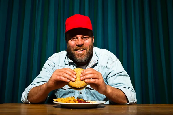 Parrakas mies syö pikaruokaa. Nautin ranskalaisista ja hampurilaisesta. Rekkakuski punaisella korkilla . — kuvapankkivalokuva