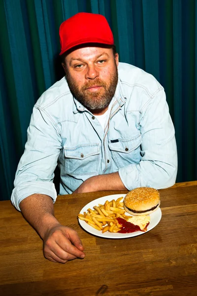 Hombre con barba comiendo comida rápida. Disfrutando de papas fritas y una hamburguesa. Camionero con gorra roja . —  Fotos de Stock