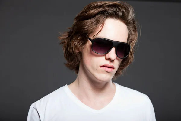 Handsome young man with brown long hair and sunglasses isolated on grey background. Fashion studio shot. Stock Picture