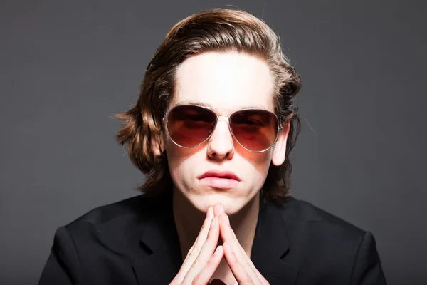 Handsome young man with brown long hair and wearing white shirt and blue jacket and sunglasses isolated on grey background. Fashion studio shot. Expressive face. Royalty Free Stock Photos