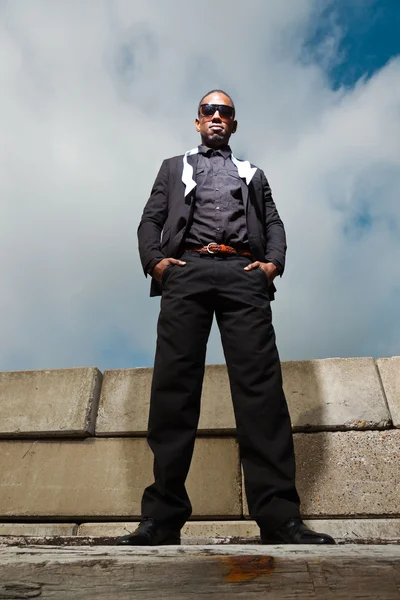 Hombre americano negro fresco en traje oscuro con gafas de sol. Fotografía de moda en entorno urbano. Cielo nublado azul . — Foto de Stock