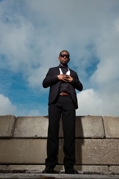 Hűvös, fekete amerikai férfi sötét öltöny napszemüveget visel. divat lövés, városi környezetben. kék felhős ég. — Stock Fotó