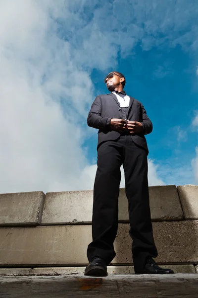 Крутой черный американец в темном костюме в солнечных очках. Мода снимается в городских условиях. Голубое облачное небо . — стоковое фото