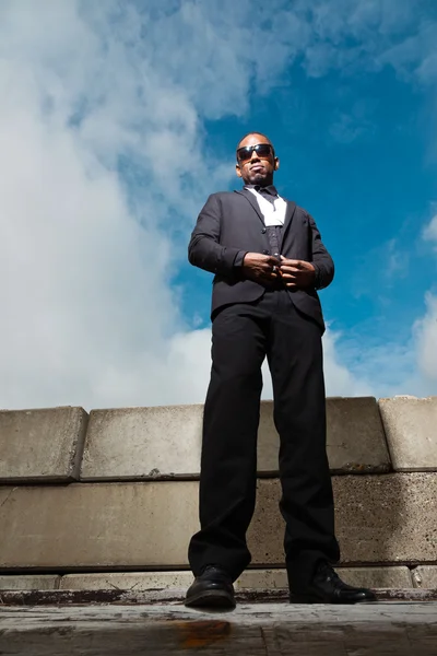 クールな黒いアメリカ スーツの男暗いサングラスを着用します。ファッション都市設定で撮影。青い曇り空. — ストック写真