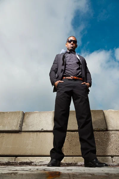Cool svart amerikansk man i mörk kostym bär solglasögon. mode skott i urban miljö. blå mulen himmel. — Stockfoto