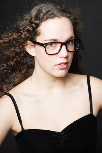Linda chica con el pelo largo y rizado marrón. Retrato de estudio de moda aislado sobre fondo negro. Vestido negro y gafas . — Foto de Stock
