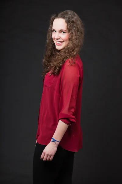 Menina bonita sorridente com cabelo encaracolado marrom longo. Retrato de estúdio de moda isolado contra fundo preto. Vestindo camisa vermelha . — Fotografia de Stock