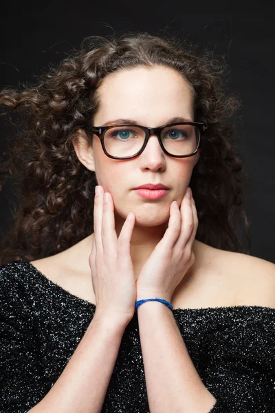 Pretty girl with long brown curly hair. Fashion studio portrait isolated against black background. Wearing black dress and glasses. — Stock Photo, Image