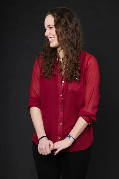 Smiling pretty girl with long brown curly hair. Fashion studio portrait isolated against black background. Wearing red shirt. — Stock Photo, Image