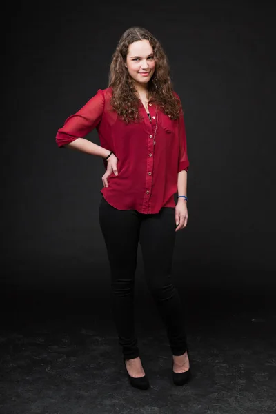 Lachende mooi meisje met lange bruin krullend haar. mode studio portret geïsoleerd tegen zwarte achtergrond. het dragen van rode shirt. — Stockfoto