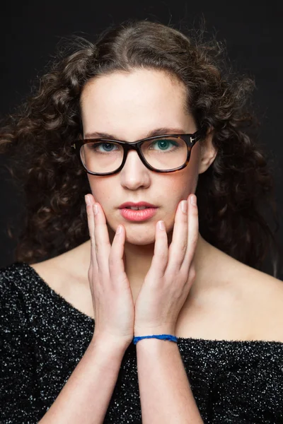 Pretty girl with long brown curly hair. Fashion studio portrait isolated against black background. Wearing black dress and glasses. — Stock Photo, Image