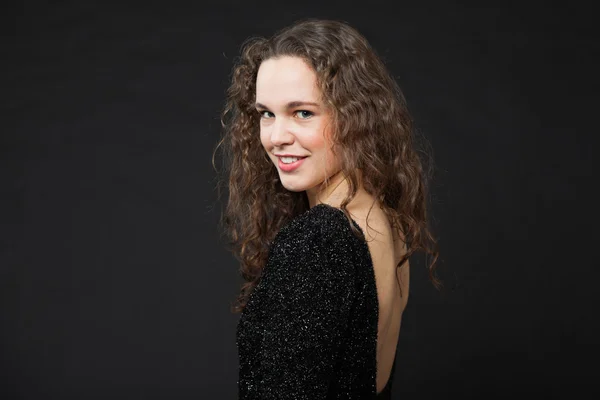 Smiling pretty girl with long brown curly hair. Fashion studio portrait isolated against black background. Wearing black dress. — Stock Photo, Image