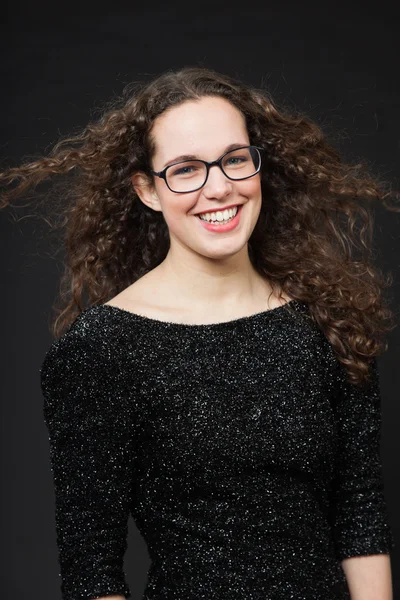 Smiling pretty girl with long brown curly hair. Fashion studio portrait isolated against black background. Wearing black dress and glasses. — Stock Photo, Image