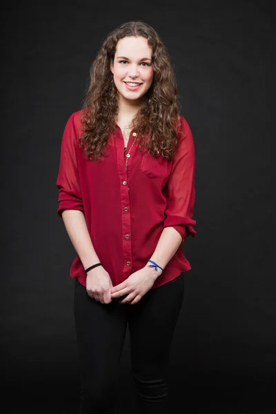Sonriente chica bonita con el pelo largo y rizado marrón. Retrato de estudio de moda aislado sobre fondo negro. Usar camisa roja . — Foto de Stock