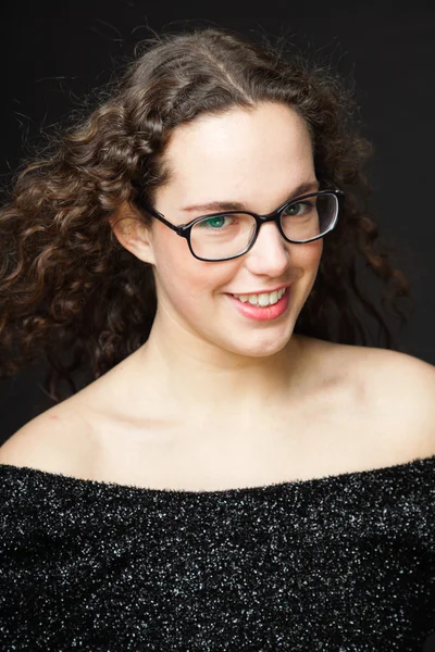 Smiling pretty girl with long brown curly hair. Fashion studio portrait isolated against black background. Wearing black dress and glasses. — Stock Photo, Image