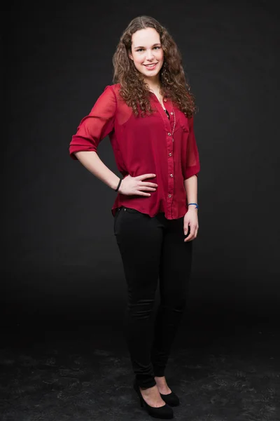 Smiling pretty girl with long brown curly hair. Fashion studio portrait isolated against black background. Wearing red shirt. — Stock Photo, Image