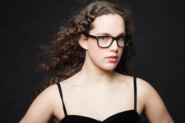 Menina bonita com cabelo encaracolado marrom longo. Retrato de estúdio de moda isolado contra fundo preto. Usando vestido preto e óculos . — Fotografia de Stock
