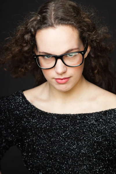 Pretty girl with long brown curly hair. Fashion studio portrait isolated against black background. Wearing black dress and glasses. Stock Photo