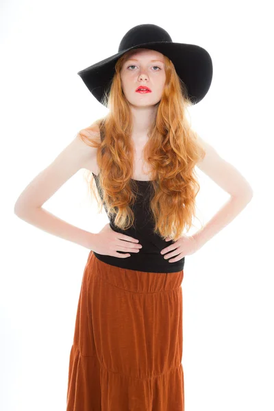 Chica guapa con el pelo largo y rojo vistiendo camisa negra y vestido marrón y sombrero negro. Estudio de moda tiro aislado sobre fondo blanco . — Foto de Stock