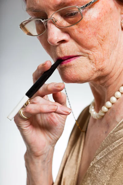 Portrait of good looking senior woman wearing glasses with expressive face showing emotions. Smoking a cigarette. Acting young. Studio shot isolated on grey background. — Stock Photo, Image