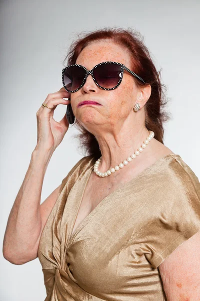 Портрет добродушної старшої жінки в сонцезахисних окулярах з виразним обличчям, що демонструє емоції. Дзвінок з мобільним телефоном. Поводитися молодо. Студійний знімок ізольовано на сірому фоні . — стокове фото