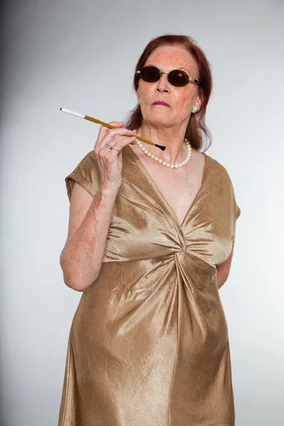 Портрет добродушної старшої жінки з виразним обличчям, що демонструє емоції. Носіння сонцезахисних окулярів і куріння цигарки. Поводитися молодо. Студійний знімок ізольовано на сірому фоні . — стокове фото