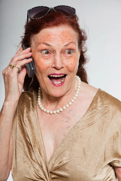 Portrait of good looking senior woman with expressive face showing emotions. Calling with cell phone. Acting young. Studio shot isolated on grey background. — Stock Photo, Image