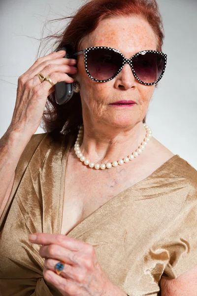 Portrait of good looking senior woman wearing sunglasses with expressive face showing emotions. Calling with cell phone. Acting young. Studio shot isolated on grey background. — Stock Photo, Image