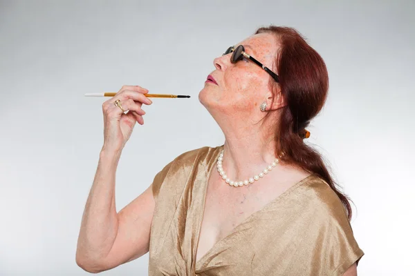 Portret van goed uitziende senior vrouw met expressief gezicht toont emoties. dragen van een zonnebril en roken van een sigaret. handelend jonge. studio opname geïsoleerd op grijze achtergrond. — Stockfoto