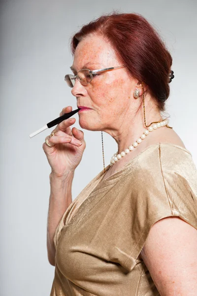 Portrait of good looking senior woman wearing glasses with expressive face showing emotions. Smoking a cigarette. Acting young. Studio shot isolated on grey background. — Stock Photo, Image