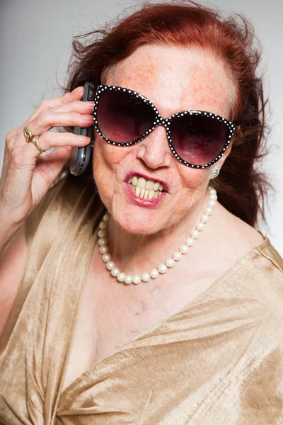 Retrato de una mujer mayor guapa usando gafas de sol con cara expresiva mostrando emociones. Llamando con el teléfono celular. Actuando joven. Estudio plano aislado sobre fondo gris . — Foto de Stock
