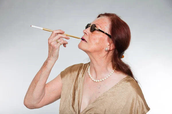 Ritratto di bella donna anziana con volto espressivo che mostra emozioni. Indossare occhiali da sole e fumare una sigaretta. Si comporta da giovane. Studio girato isolato su sfondo grigio . — Foto Stock