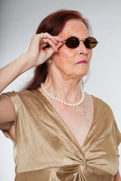 Portrait of good looking senior woman wearing sunglasses with expressive face showing emotions. Acting young. Studio shot isolated on grey background. — Stock Photo, Image