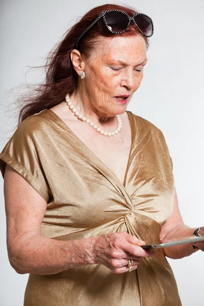 Retrato de mulher idosa de boa aparência usando óculos de sol com rosto expressivo mostrando emoções. Usando um tablet. A agir jovem. Estúdio tiro isolado em fundo cinza . — Fotografia de Stock
