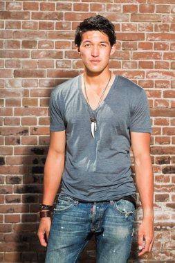 Urban asian man. Good looking. Cool guy. Wearing grey shirt and jeans. Standing in front of brick wall. clipart