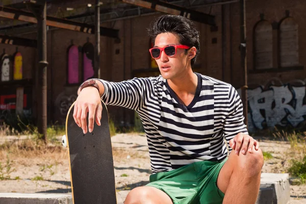 Urbain asiatique homme avec des lunettes de soleil rouges et skateboard assis sur la rue. Beau look. Cool mec. Portant un pull rayé bleu blanc et un short vert. Ancien bâtiment négligé en arrière-plan . — Photo