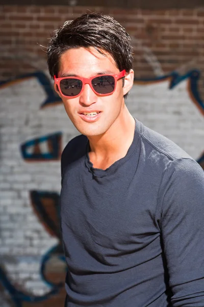 Stedelijke Aziatische man met rode zonnebril. Knap. koele kerel. het dragen van donker blauw shirt. staande voor bakstenen muur met graffiti. — Stockfoto