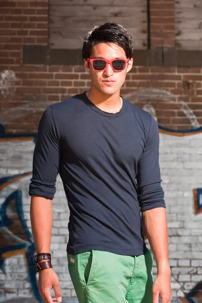 Stedelijke Aziatische man met rode zonnebril. Knap. koele kerel. dragen donkere blauwe shirt en groene shorts. staande voor bakstenen muur met graffiti. — Stockfoto