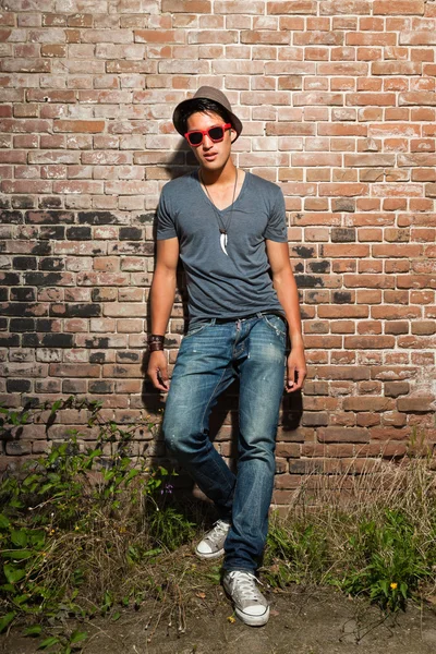 Городской азиат в красных солнечных очках. Симпатичный. Крутой парень. В серой рубашке, шляпе и джинсах. Стоя перед кирпичной стеной . — стоковое фото