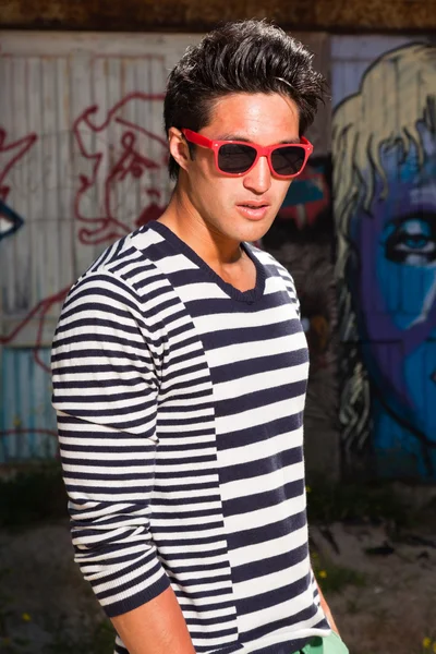 Hombre asiático urbano con gafas de sol rojas. Muy guapo. Un tipo genial. Usando jersey azul a rayas blancas. De pie frente a la pared de madera con graffiti . — Foto de Stock